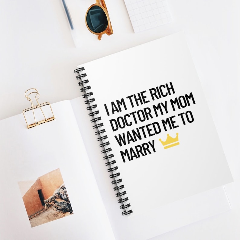 May include: A white spiral notebook with the text "I AM THE RICH DOCTOR MY MOM WANTED ME TO MARRY" in black text and a gold crown icon.