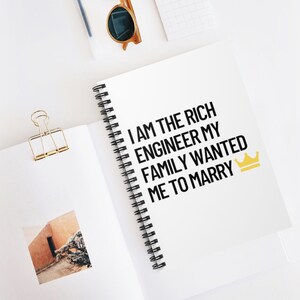Pode incluir: Um caderno branco em espiral com o texto "I am the rich engineer my family wanted me to marry" em texto preto e um ícone de coroa dourada.