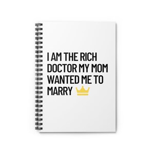 May include: White spiral notebook with the text "I AM THE RICH DOCTOR MY MOM WANTED ME TO MARRY" in black text and a gold crown.