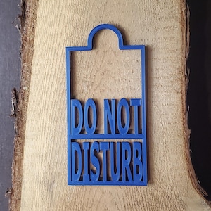 May include: A blue rectangular door hanger with a rounded top cutout. The sign features the text DO NOT DISTURB in raised, bold, capital letters. The design is constructed from a single piece of blue material with a clean, structured frame.