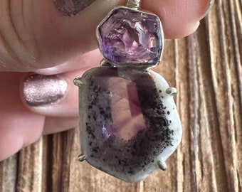 May include: A silver pendant with two amethyst stones. The top stone is a deep purple and the bottom stone is a lighter purple with white inclusions. The pendant has a decorative bail.