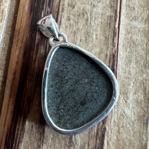 May include: A silver pendant with a dark gray stone set in a teardrop shape. The pendant has a bail on the top and the number "925" is engraved on the back.