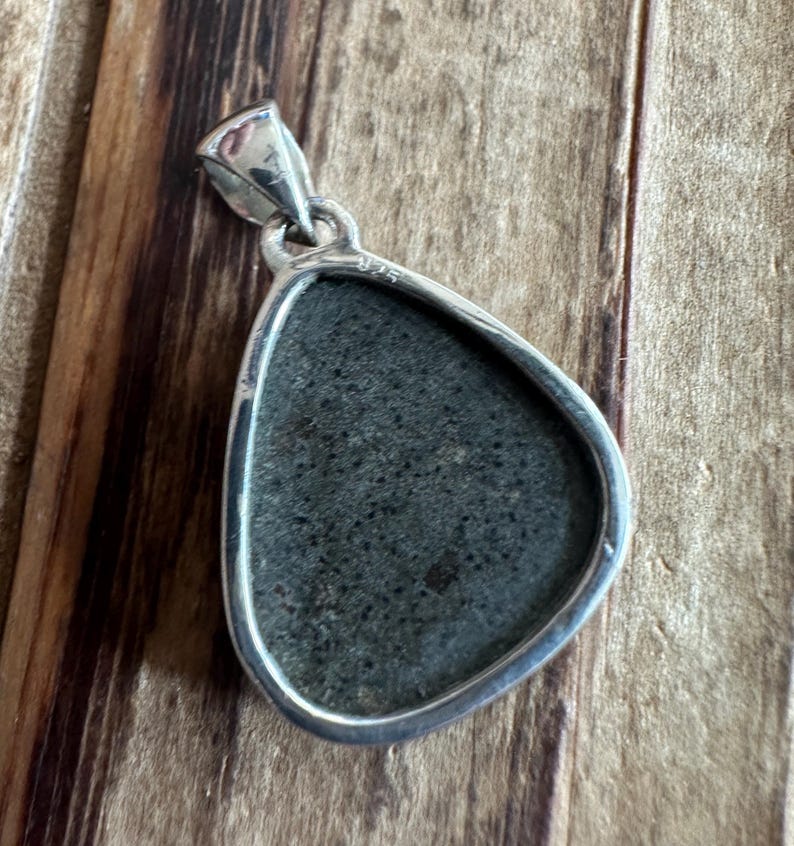 May include: A silver pendant with a dark gray stone set in a teardrop shape. The pendant has a bail on the top and the number "925" is engraved on the back.