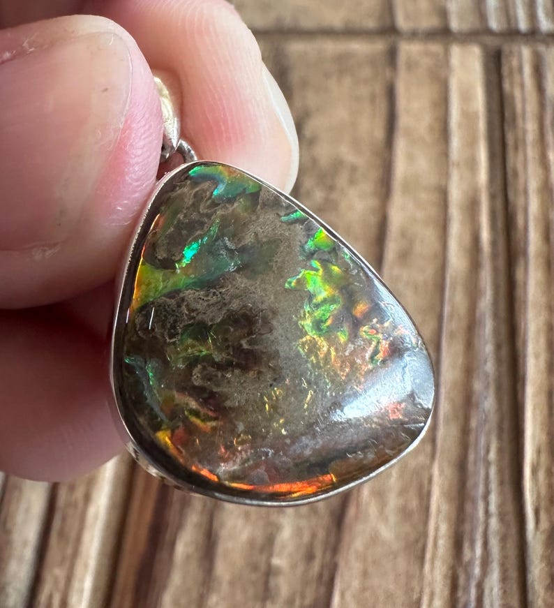 May include: A silver pendant with a teardrop-shaped iridescent gemstone. The gemstone has a dark brown base with flashes of green, blue, and orange.