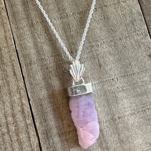 Puede incluir: Un collar de cadena de plata con un colgante que presenta un cristal crudo de color p&uacute;rpura claro. El cristal est&aacute; engastado en un bisel de plata y cuelga de una argolla de plata con un dise&ntilde;o decorativo.