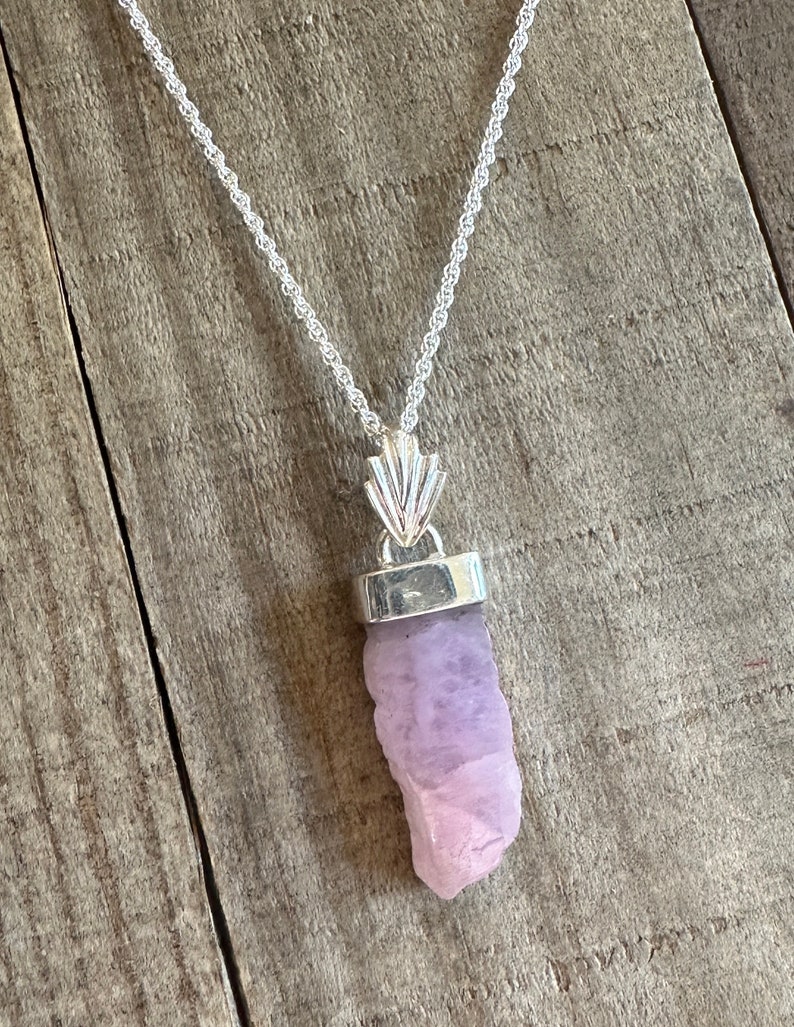 Puede incluir: Un collar de cadena de plata con un colgante que presenta un cristal crudo de color p&uacute;rpura claro. El cristal est&aacute; engastado en un bisel de plata y cuelga de una argolla de plata con un dise&ntilde;o decorativo.