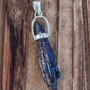 May include: A silver pendant with a dark iridescent crystal. The crystal is shaped like a long, thin point and has a metallic sheen. The pendant is on a wooden background.