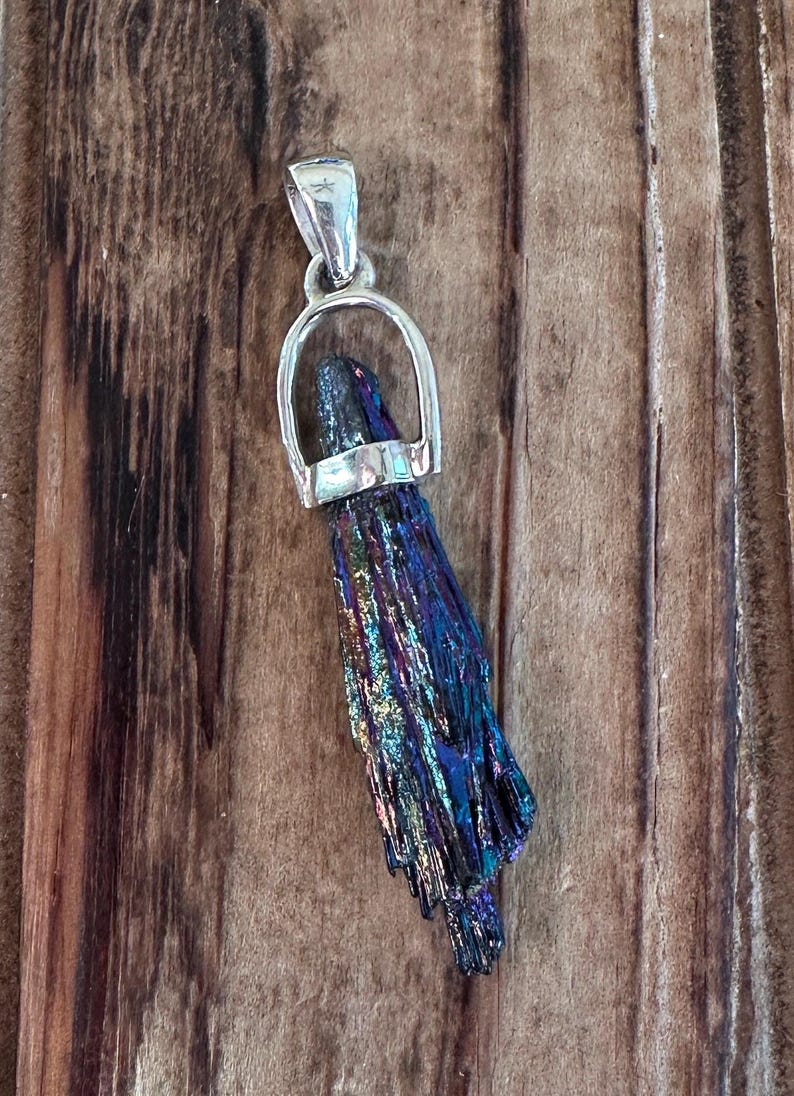 May include: A silver pendant with a dark iridescent crystal. The crystal is shaped like a long, thin point and has a metallic sheen. The pendant is on a wooden background.