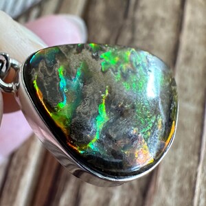 May include: A silver pendant with a teardrop-shaped iridescent gemstone. The gemstone has a dark gray base with green, blue, and orange iridescent flashes.