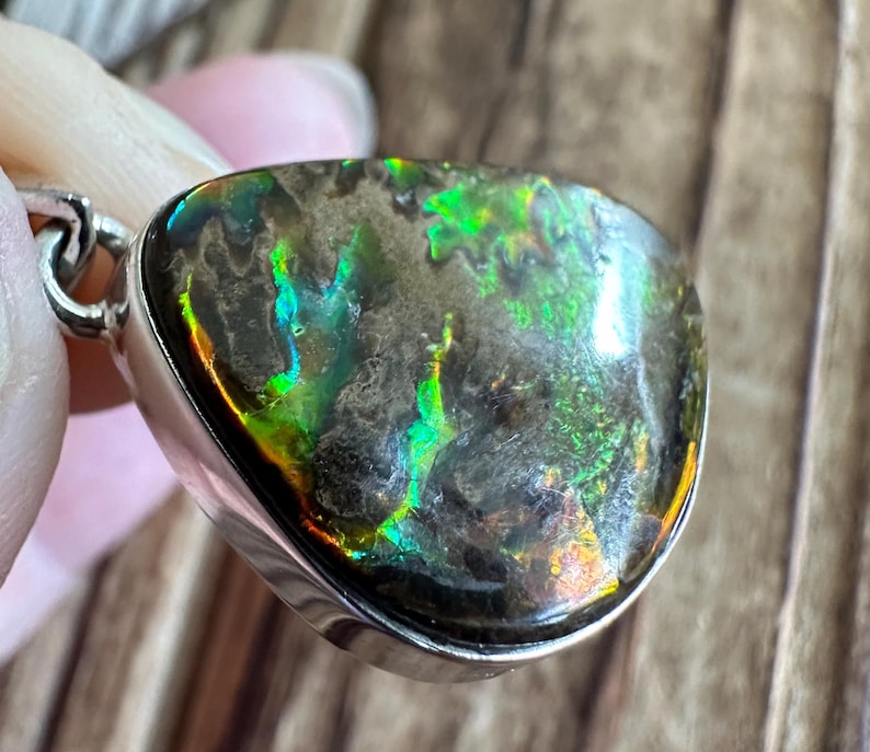 May include: A silver pendant with a teardrop-shaped iridescent gemstone. The gemstone has a dark gray base with green, blue, and orange iridescent flashes.