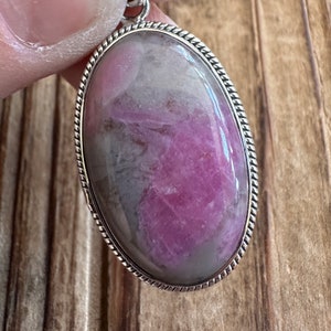 May include: A silver pendant with a pink and gray oval gemstone set in a rope-like border.