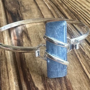 May include: A silver bangle bracelet featuring a rectangular, blue gemstone centerpiece. The gemstone is wrapped with silver bands, creating a unique design. The bracelet is displayed on a wooden surface.