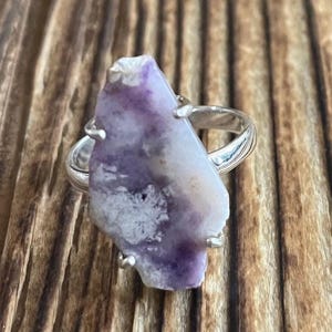 May include: A silver ring with a large, irregular shaped purple and white stone set in the center.