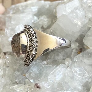 Pu&ograve; includere: Un anello d'argento con una grande pietra trasparente di forma ovale. La pietra presenta inclusioni interne ed &egrave; circondata da un motivo a catena. La fascia dell'anello &egrave; larga e lucida. L'anello &egrave; posto su uno sfondo di cristalli trasparenti.