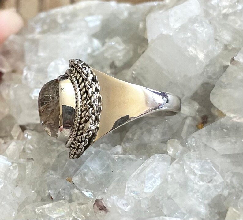 Pu&ograve; includere: Un anello d'argento con una grande pietra trasparente di forma ovale. La pietra presenta inclusioni interne ed &egrave; circondata da un motivo a catena. La fascia dell'anello &egrave; larga e lucida. L'anello &egrave; posto su uno sfondo di cristalli trasparenti.