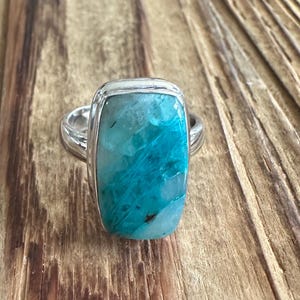 May include: A silver ring featuring a rectangular, polished gemstone with vibrant turquoise and teal marbling, set in a smooth metal bezel. The stone displays natural inclusions and a glossy surface, mounted on a simple, polished silver band.