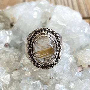 Pu&ograve; includere: Anello d'argento con una gemma ovale e trasparente contenente inclusioni dorate. L'anello ha un bordo intrecciato dettagliato e una fascia pi&ugrave; ampia. L'anello &egrave; esposto su un letto di cristalli trasparenti.