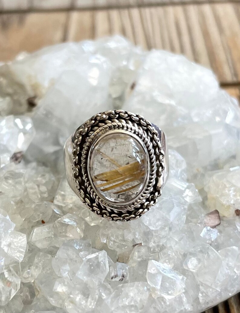 Pu&ograve; includere: Anello d'argento con una gemma ovale e trasparente contenente inclusioni dorate. L'anello ha un bordo intrecciato dettagliato e una fascia pi&ugrave; ampia. L'anello &egrave; esposto su un letto di cristalli trasparenti.