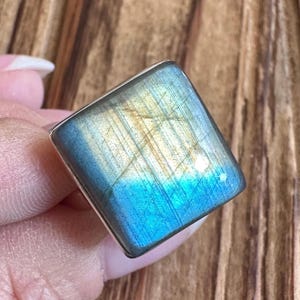 May include: Square-shaped labradorite ring with a silver band. The gemstone displays a vibrant play of color, with hues of blue, gold, and green. The ring is held in a hand, showcasing the polished surface and unique patterns of the stone.