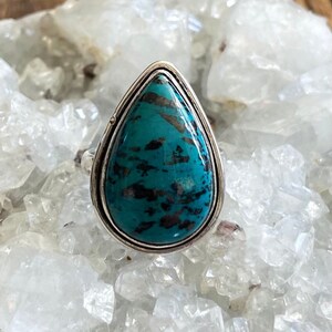 May include: A teardrop-shaped turquoise ring set in sterling silver. The turquoise is a deep blue-green color with black veins running through it.