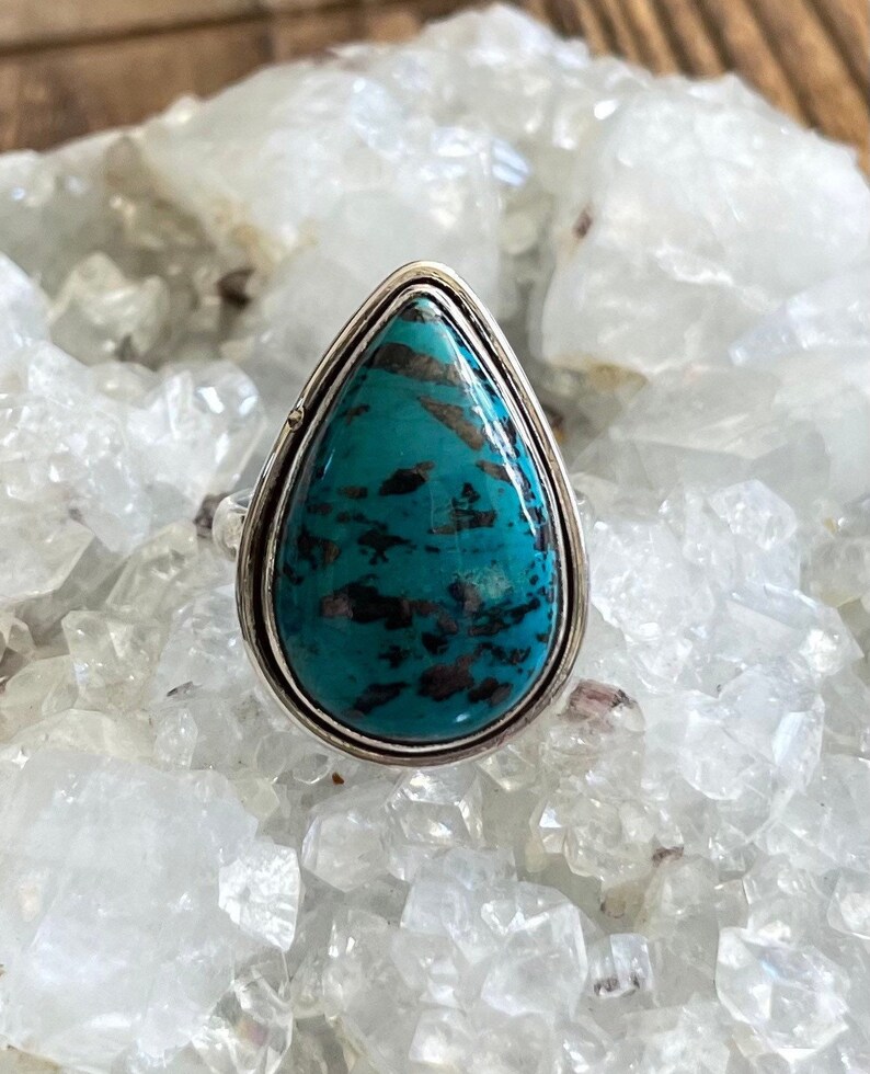 May include: A teardrop-shaped turquoise ring set in sterling silver. The turquoise is a deep blue-green color with black veins running through it.