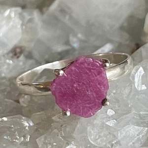 May include: A silver ring with a pink, rough-cut gemstone. The ring is set on a white background.