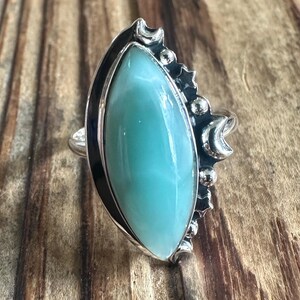 May include: A silver ring featuring a large, marquise-cut, light blue Larimar stone. The metal setting is decorated with small silver spheres, star shapes, and crescent moon motifs along one side, creating a detailed celestial-inspired design.
