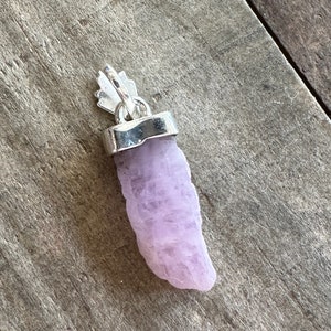 Puede incluir: Un colgante de cristal de color lavanda p&aacute;lido con una argolla de plata. El cristal es rugoso y tiene una forma natural.