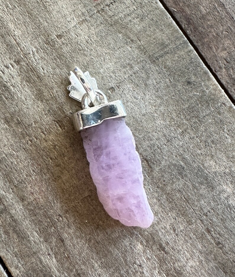 Puede incluir: Un colgante de cristal de color lavanda p&aacute;lido con una argolla de plata. El cristal es rugoso y tiene una forma natural.