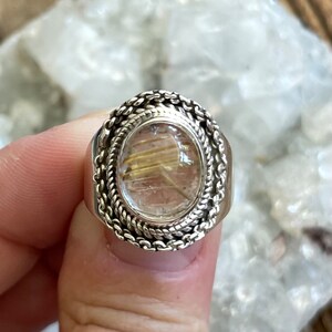 Pu&ograve; includere: Anello ovale in argento con una pietra trasparente color oro. La pietra presenta inclusioni dorate. L'anello ha un design a corda intrecciata attorno alla pietra e una fascia spessa.