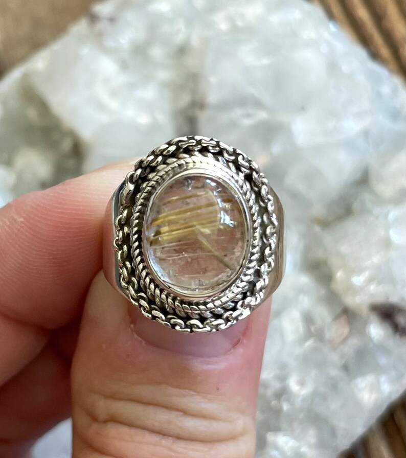 Pu&ograve; includere: Anello ovale in argento con una pietra trasparente color oro. La pietra presenta inclusioni dorate. L'anello ha un design a corda intrecciata attorno alla pietra e una fascia spessa.