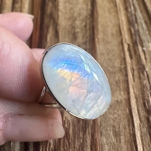 May include: A silver ring featuring a large, oval-shaped moonstone cabochon. The stone displays a translucent white appearance with iridescent blue and orange flashes of light across its smooth, polished surface, set within a simple metal band.