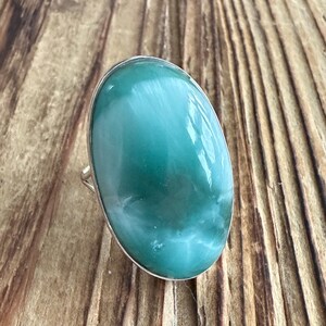 May include: A large oval ring featuring a polished, light teal gemstone with white marbling. The gemstone is set in a silver band. The ring is displayed on a wooden surface.