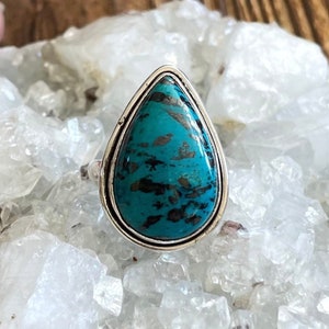 May include: A teardrop-shaped turquoise ring set in sterling silver. The turquoise is a deep blue-green color with black and brown veins.