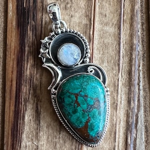 May include: A silver pendant with a turquoise gemstone and a moonstone. The pendant is intricately designed with a silver frame and a beaded border.