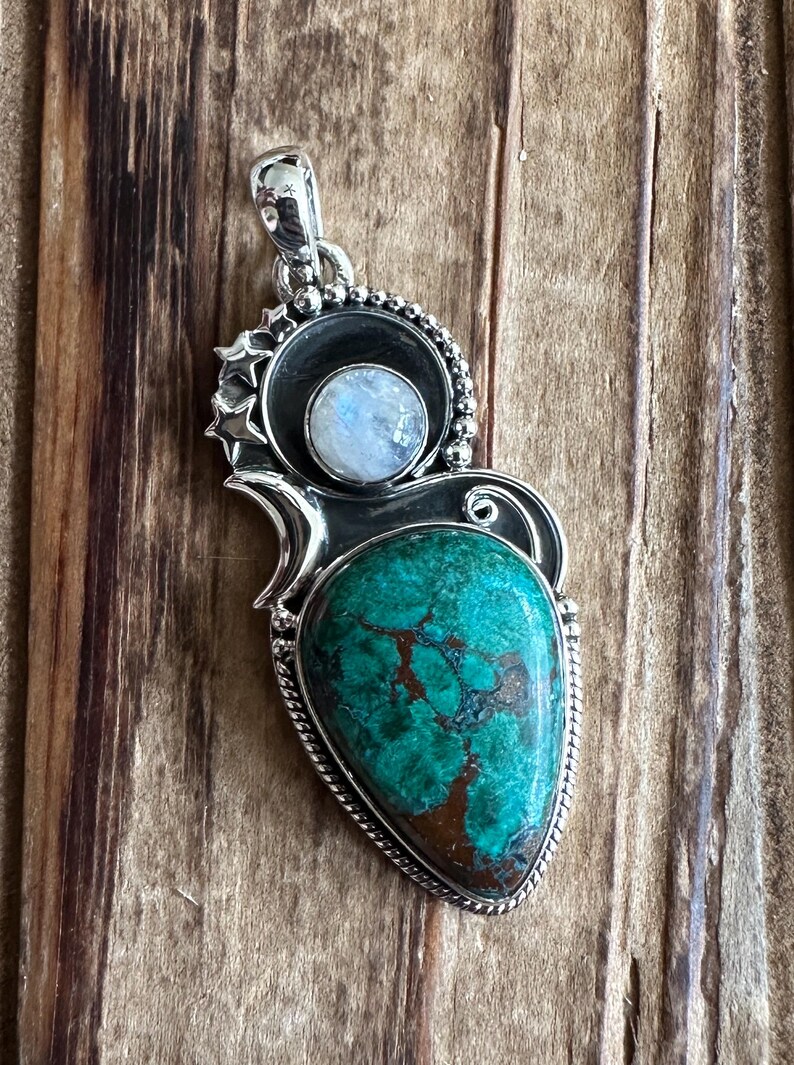May include: A silver pendant with a turquoise gemstone and a moonstone. The pendant is intricately designed with a silver frame and a beaded border.