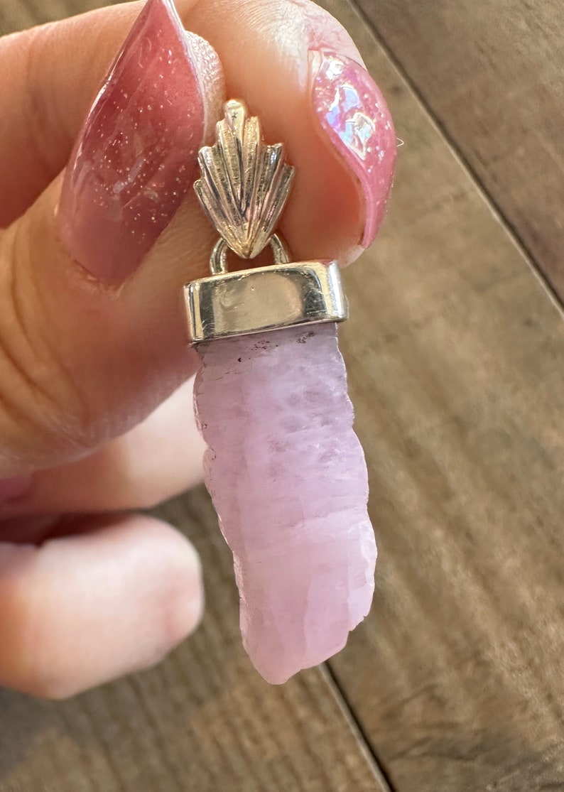 Puede incluir: Un colgante de plata con un cristal rosa. El cristal es de forma natural y tiene un color rosa claro.