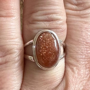 May include: A silver ring with a large oval-shaped sunstone. The sunstone is a deep orange color with flecks of gold.