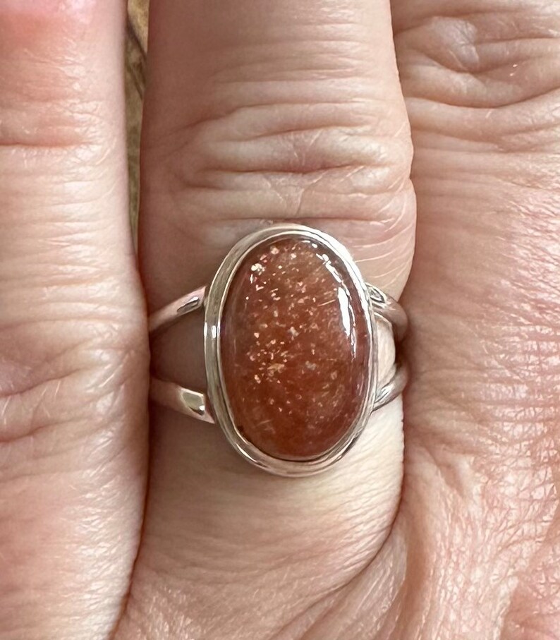 May include: A silver ring with a large oval-shaped sunstone. The sunstone is a deep orange color with flecks of gold.