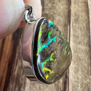 May include: A silver pendant with a teardrop-shaped opal stone. The opal has a dark gray base with iridescent green, blue, and yellow flashes.