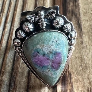 May include: A silver ring featuring a green and pink gemstone, with a bee design on the top.
