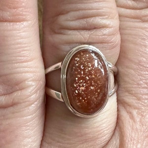 May include: A silver ring with a large oval-shaped sunstone. The sunstone is a reddish-brown color with gold flecks.