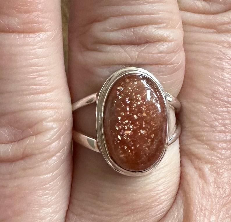 May include: A silver ring with a large oval-shaped sunstone. The sunstone is a reddish-brown color with gold flecks.