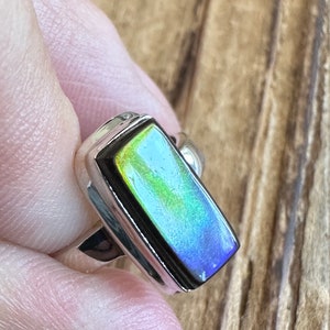 K&ouml;nnte beinhalten: Ein silberner Ring mit einem rechteckigen, schillernden Edelstein, der in einer Zargenfassung sitzt. Der Edelstein zeigt eine Reihe von Farben, darunter Gr&uuml;n, Blau und Gelb.