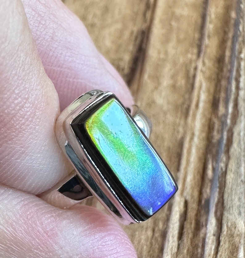 K&ouml;nnte beinhalten: Ein silberner Ring mit einem rechteckigen, schillernden Edelstein, der in einer Zargenfassung sitzt. Der Edelstein zeigt eine Reihe von Farben, darunter Gr&uuml;n, Blau und Gelb.