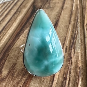 May include: A teardrop-shaped ring featuring a large, polished larimar stone. The stone displays a mix of light blue and turquoise hues with white marbling, set in a silver band. The ring's design is simple, highlighting the natural beauty of the gemstone.