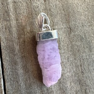 Puede incluir: Un colgante de cristal de color lavanda p&aacute;lido con una argolla de plata. El cristal tiene una forma natural y toscamente cortada.
