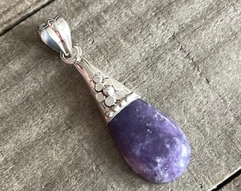 May include: A teardrop-shaped pendant with a polished, deep purple stone. The pendant has a silver-colored metal setting with a decorative floral design at the top. The stone is approximately 1.5 inches long.