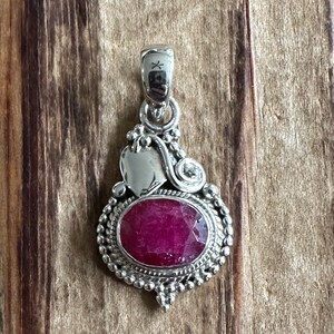May include: A silver pendant with a red gemstone set in a detailed silver frame. The pendant has a heart-shaped design on the top and a small silver bead on the side.
