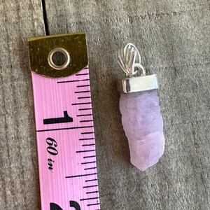 Puede incluir: Un colgante de cristal de color lavanda p&aacute;lido con una anilla de plata. El cristal mide aproximadamente 2,5 cm de largo y tiene una textura rugosa y natural. El colgante se muestra junto a una cinta m&eacute;trica rosa.